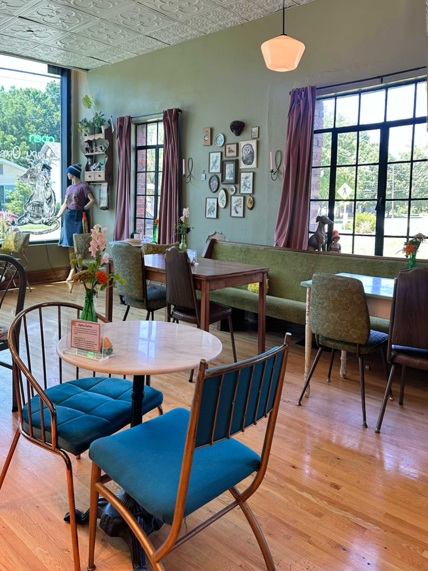 Inviting local coffee shop interior featuring vintage furnishings, colorful chairs, wood floors, floral arrangements, and large sunlit windows.