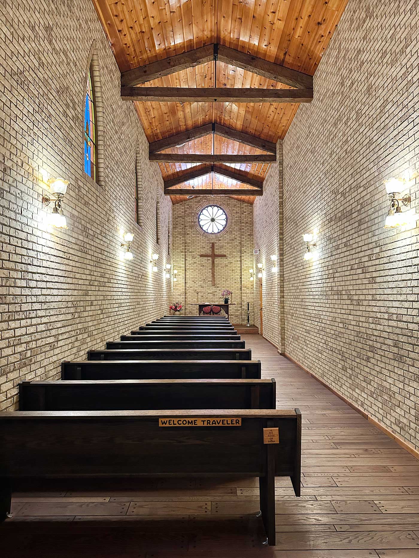 Wooden pews inside a narrow chapel with white brick walls and a vaulted wood ceiling.