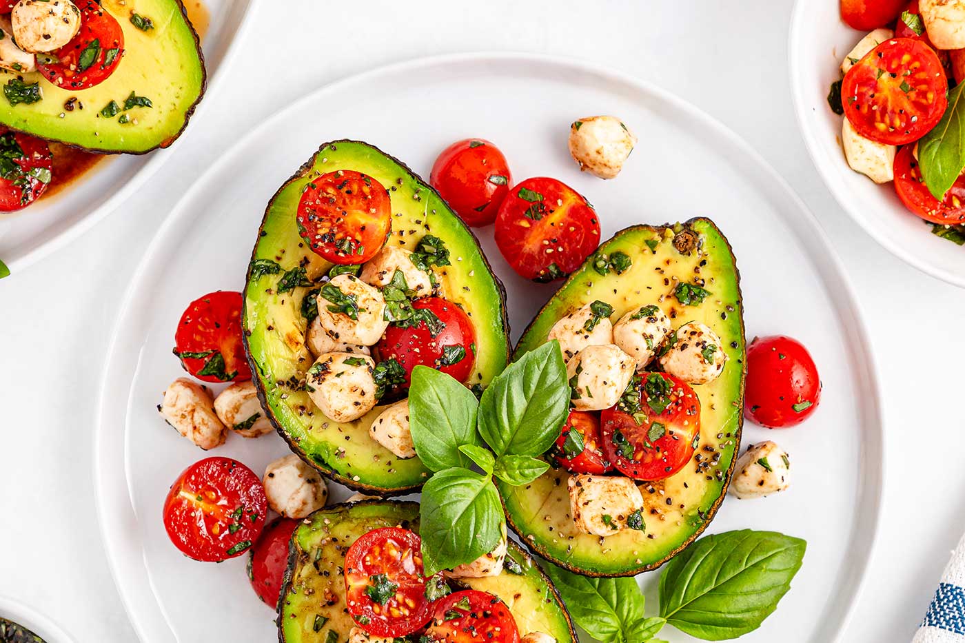 Caprese stuffed avocado halves topped with cherry tomatoes, mozzarella pearls, fresh basil, and black pepper on a white plate.