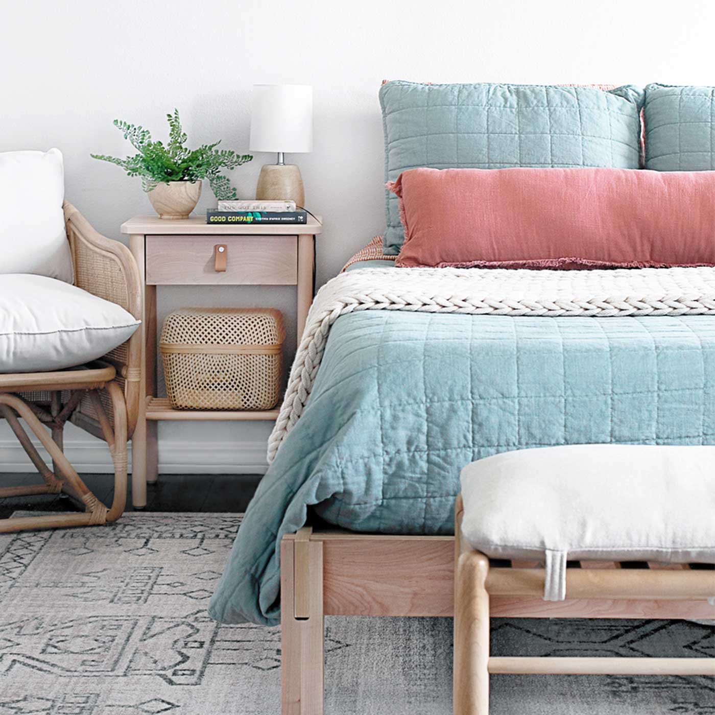 Bright master bedroom with soft blue bedding, a coral lumbar pillow, woven furniture, and a light wood nightstand.