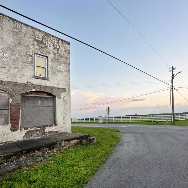 Weathered brick and stucco building beside a quiet country road with green grass, white fencing, utility lines, and soft pink clouds at sunset.