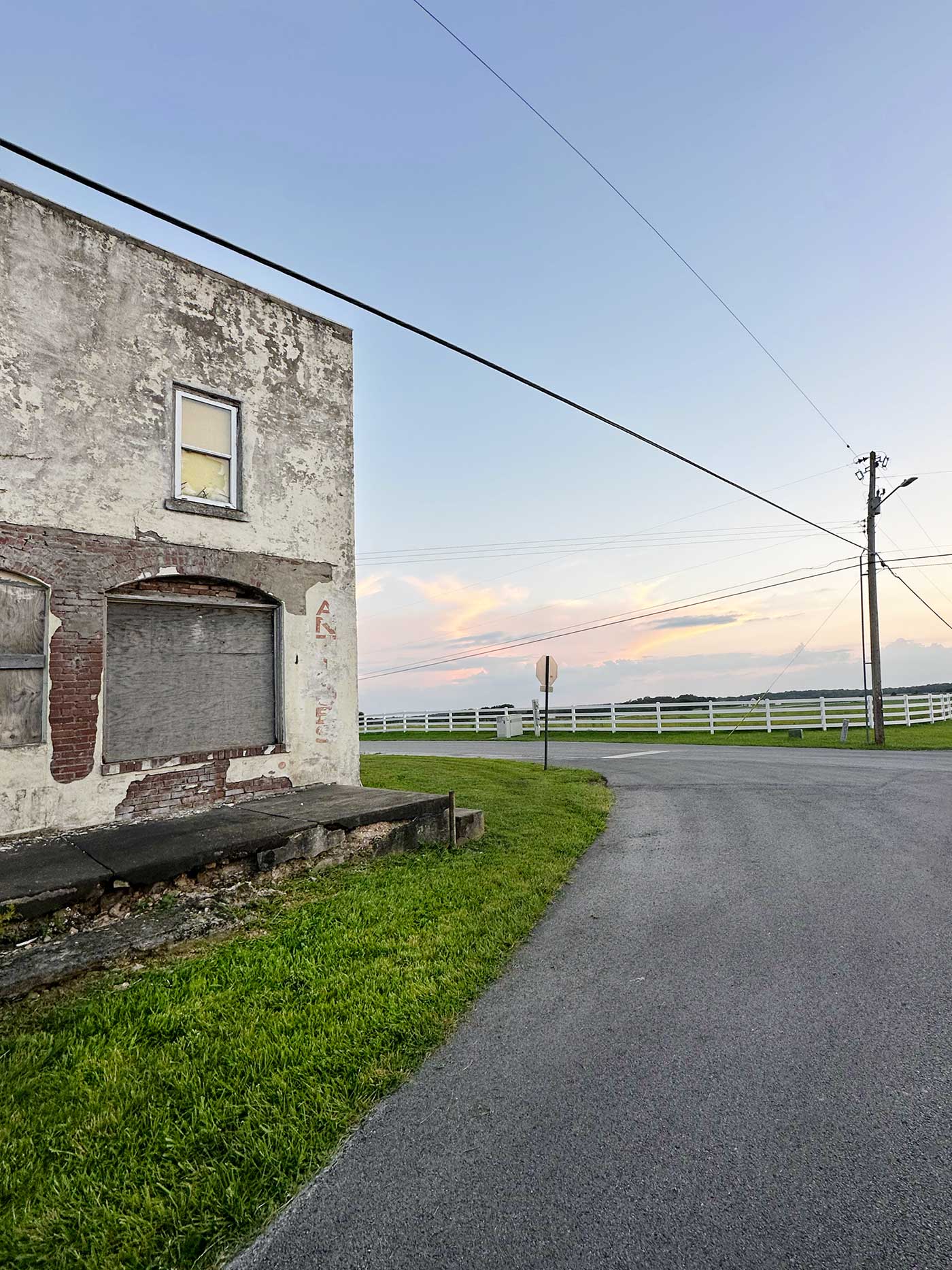Weathered old brick and stucco building beside a quiet country road with white fencing and a pink-and-gold sunset in the distance.