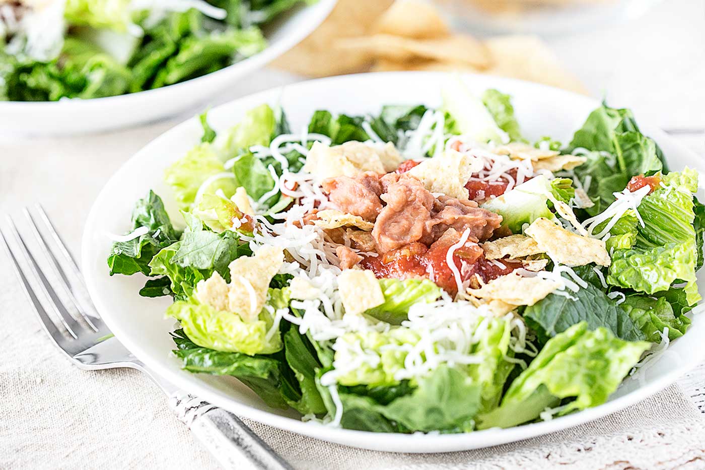 Taco salad with romaine lettuce, refried beans, salsa, shredded cheese, and crushed tortilla chips in a white bowl.