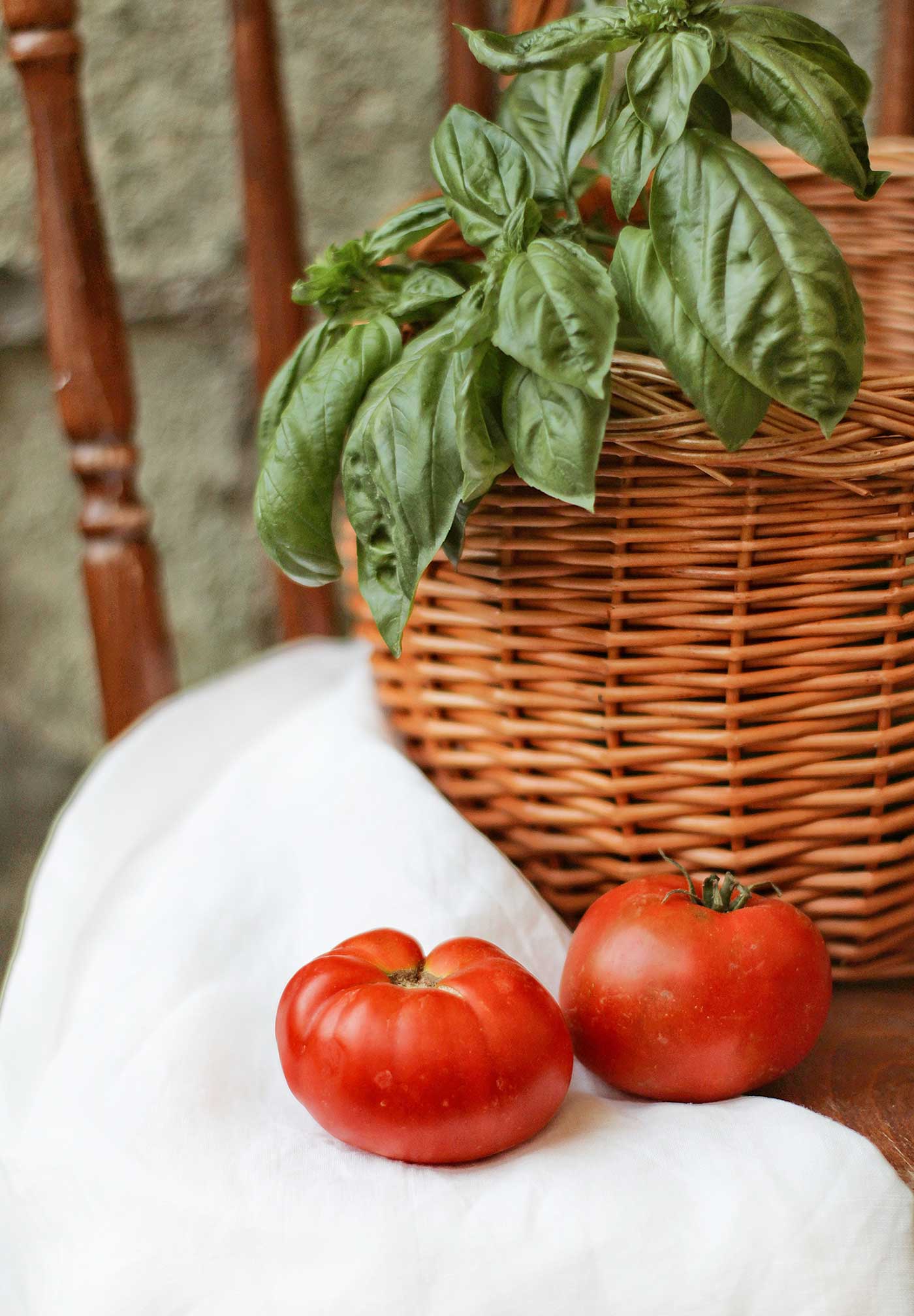 Two ripe red tomatoes beside a potted basil plant in a woven basket.