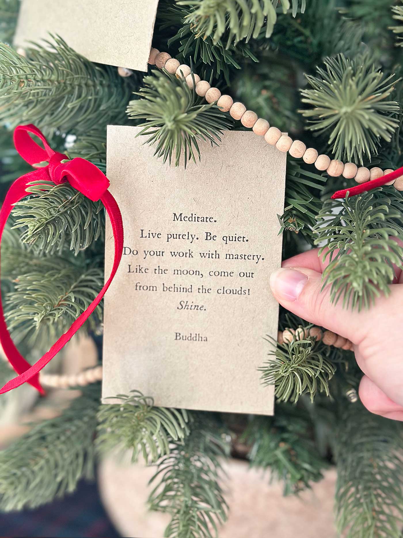 A hand placing a quote card onto a tabletop Christmas tree decorated with wooden bead garland and red velvet ribbon.
