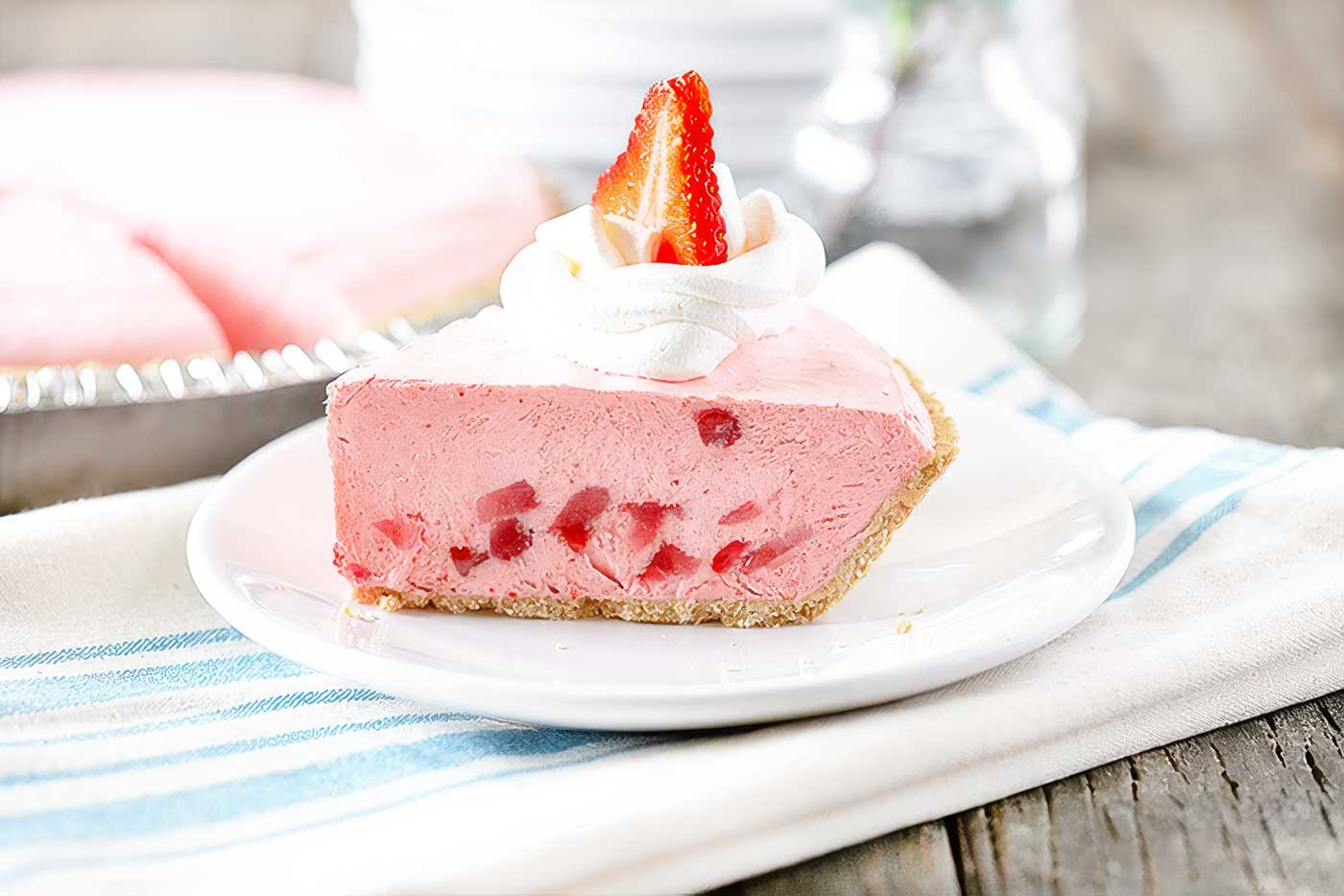 Slice of no-bake strawberry pie with a graham cracker crust, whipped cream, and a fresh strawberry on a white plate.
