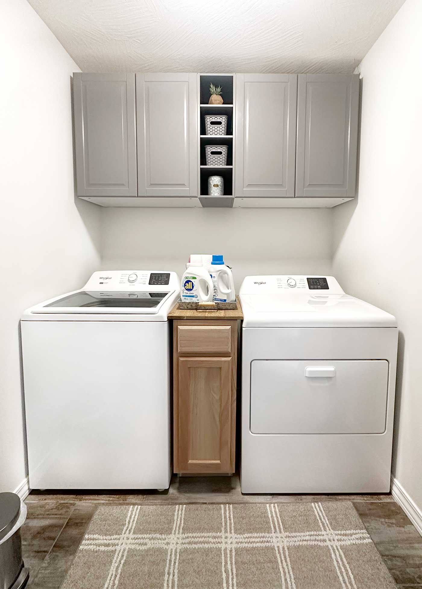 Bright, organized laundry room featuring a new white top-load washer, matching dryer, gray cabinets, woven baskets, and a neutral plaid rug.
