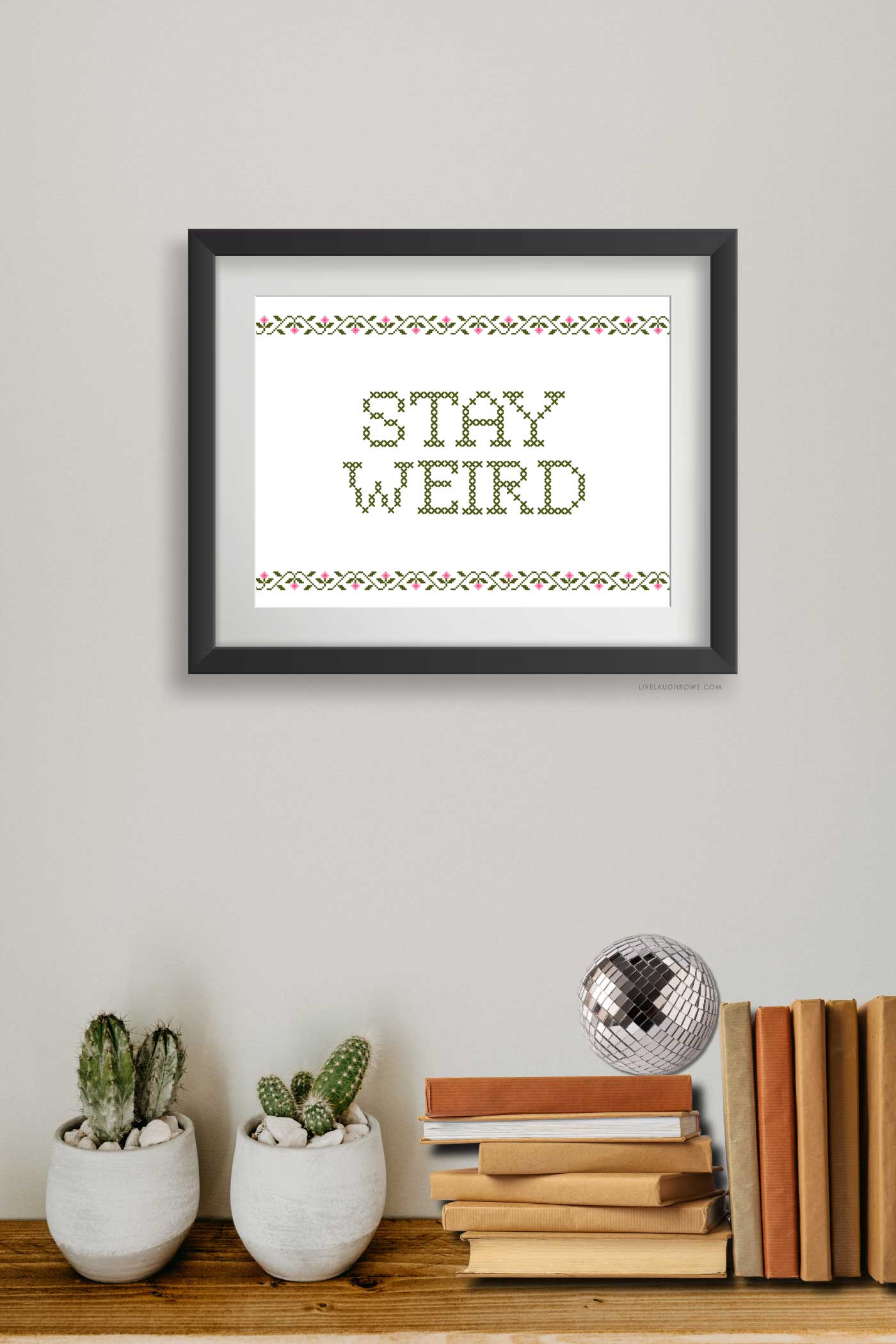 Framed Stay Weird printable displayed above potted cacti, stacked books, and a small disco ball