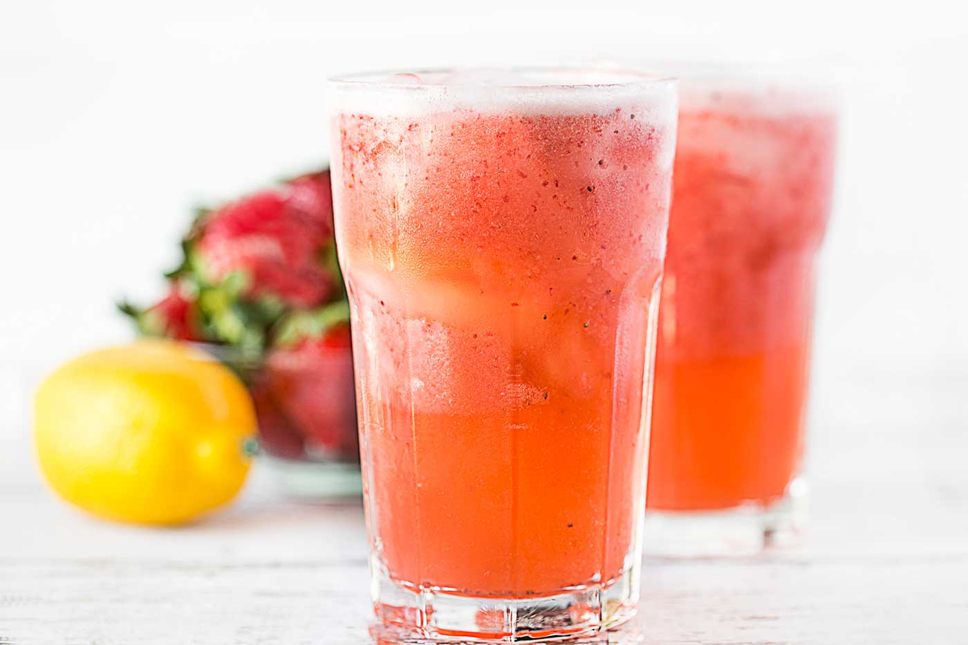 Two glasses of strawberry lemonade cooler served over ice with fresh strawberries and a lemon in the background.