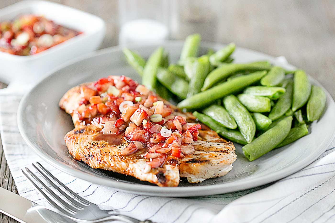 Grilled chicken topped with fresh strawberry salsa and served with sugar snap peas on a gray plate.
