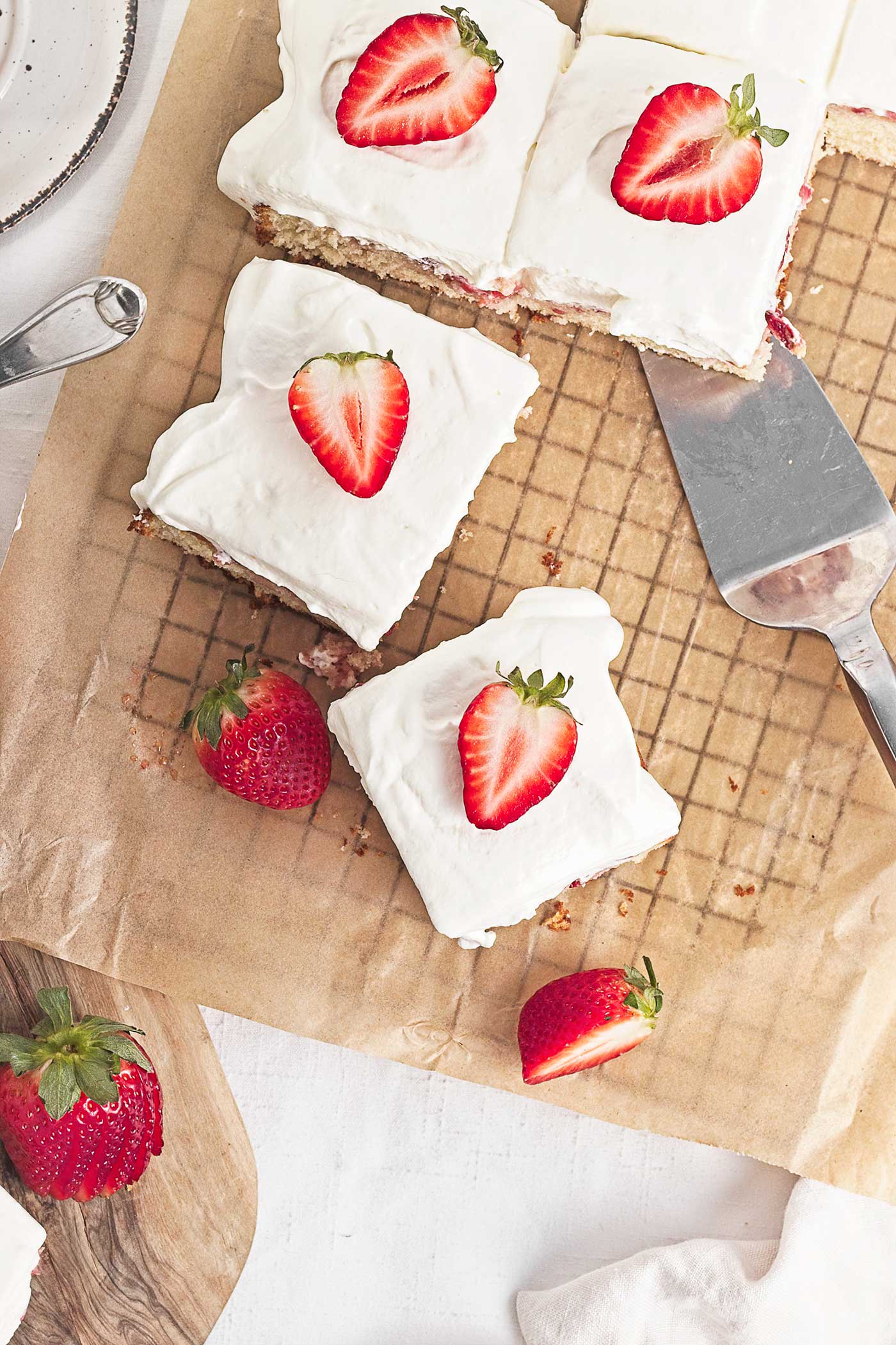 Strawberry shortcake sheet cake squares with a cake server and fresh strawberries on parchment paper