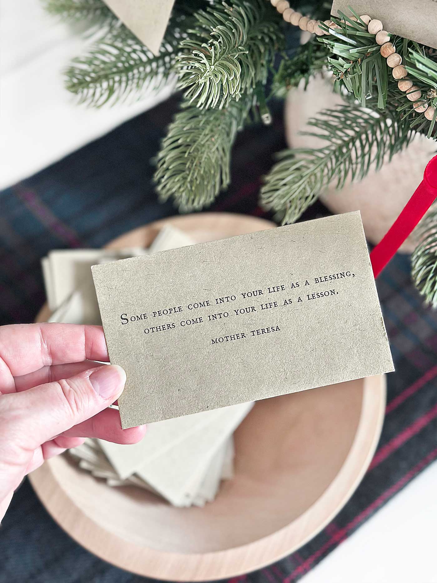 A hand holding a quote card above a bowl filled with additional quote cards beside a tabletop Christmas tree.