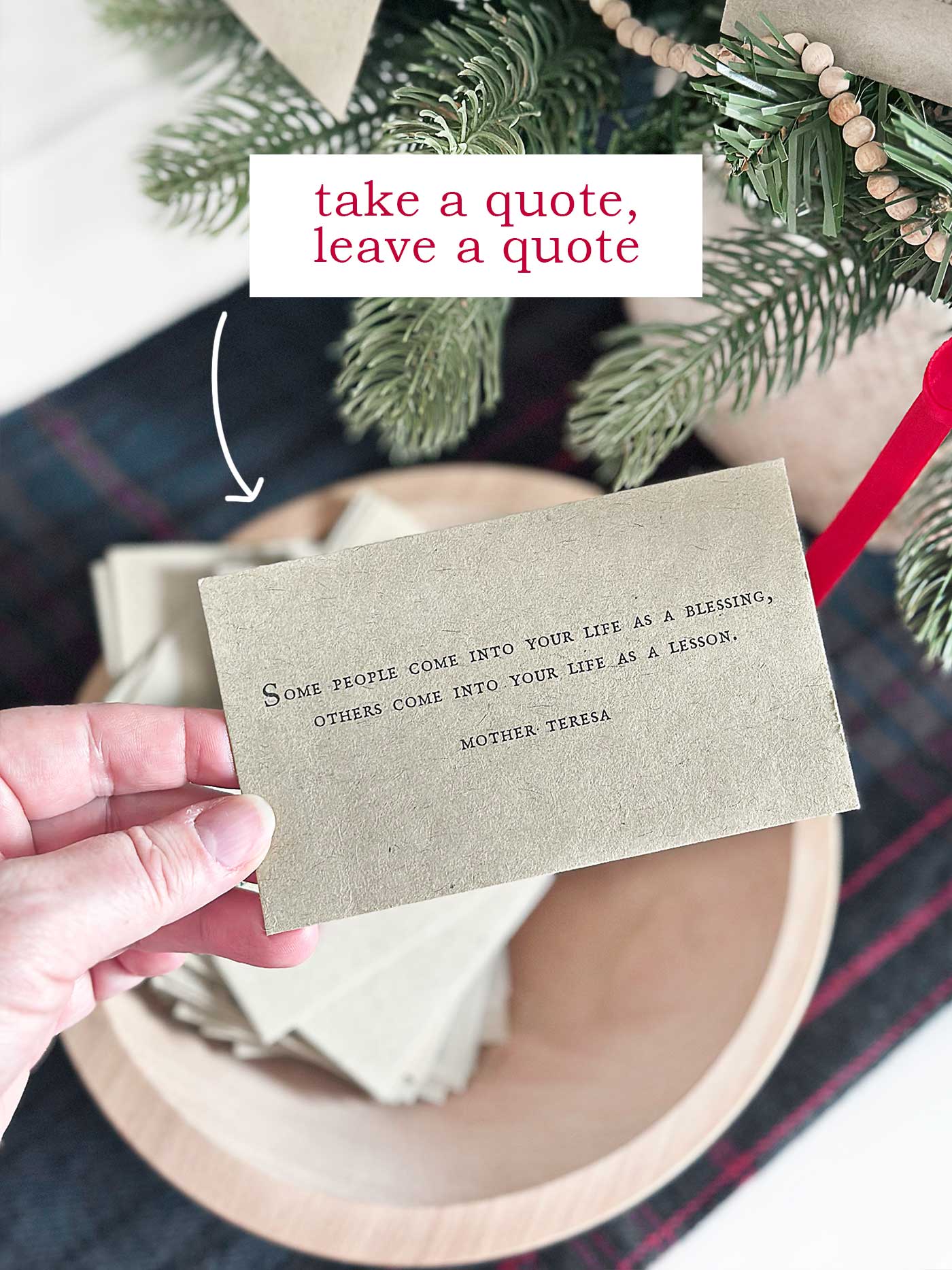 A hand holding a quote card above a bowl filled with additional quote cards beside a tabletop Christmas tree with text overlay, take a quote, leave a quote.