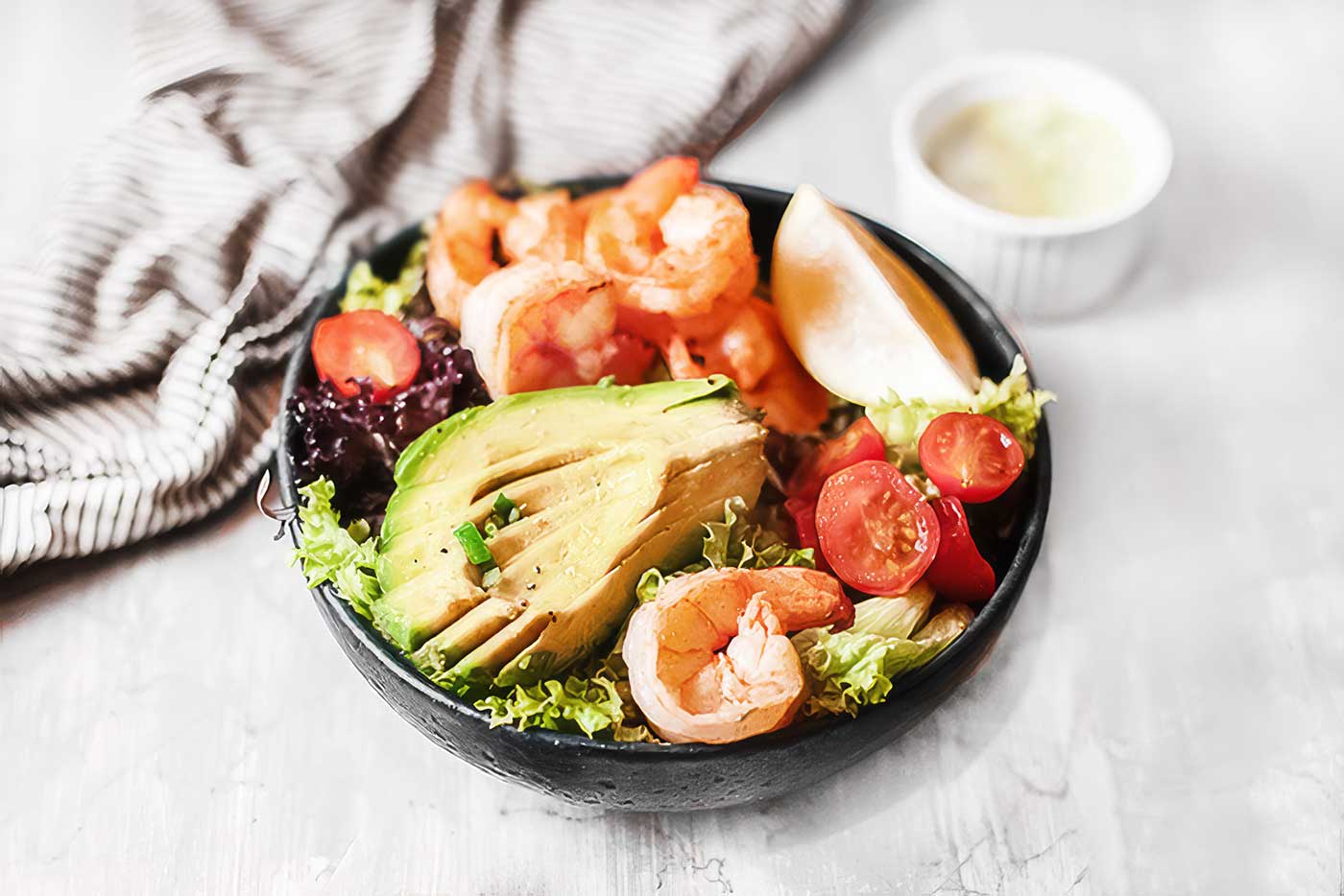 Zesty shrimp avocado salad with leafy greens, cherry tomatoes, sliced avocado, and lemon in a black bowl. Easy Summer Recipe.