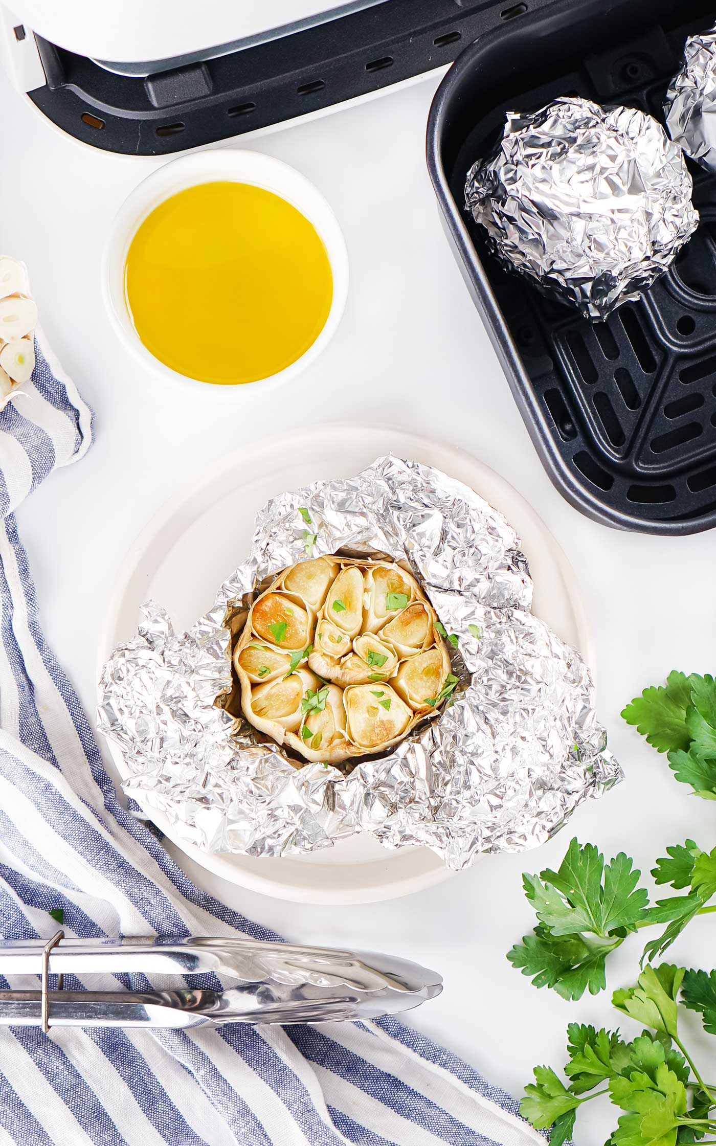 Roasted garlic bulb resting in opened aluminum foil beside an air fryer and olive oil.
