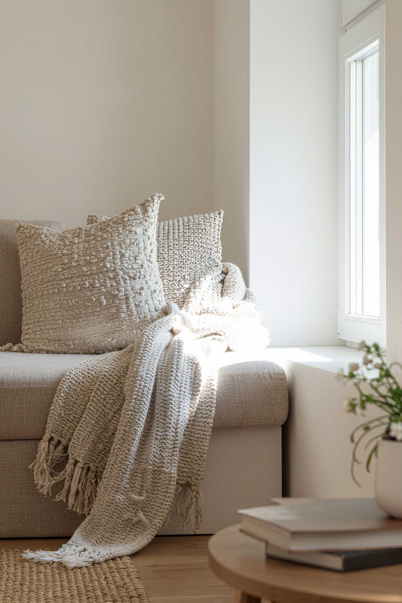 Cozy neutral chair by a sunny window with textured cream pillows, a chunky knit throw, books on a round table, and a soft woven rug.