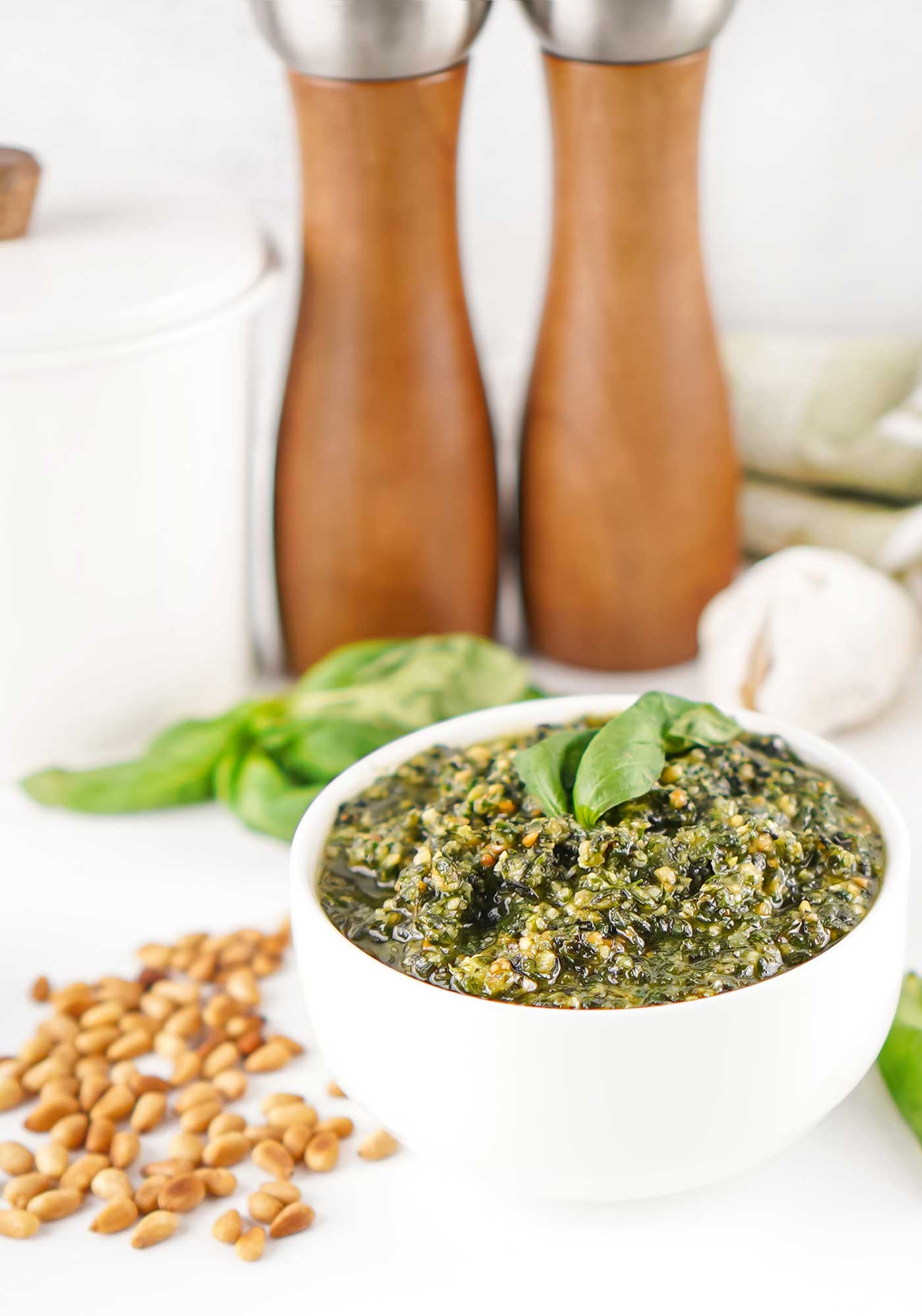 Homemade basil pesto in a white bowl with fresh basil, garlic, pine nuts, and wooden salt and pepper mills in the background.