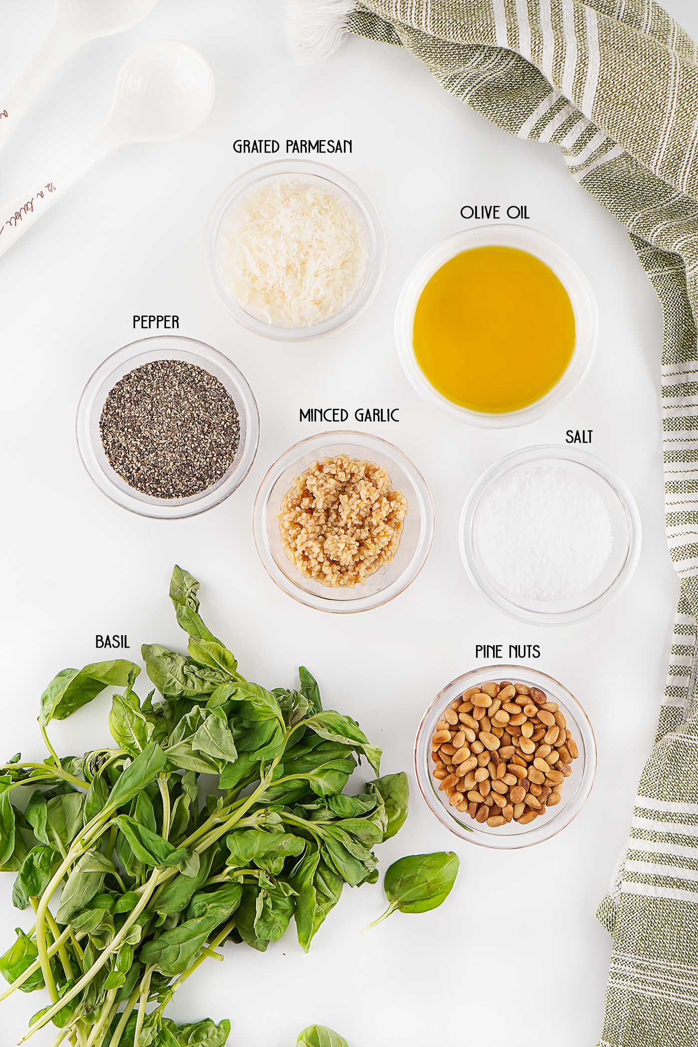 Ingredients for pesto including fresh basil, olive oil, Parmesan cheese, garlic, pine nuts, salt, and pepper arranged on a white surface.