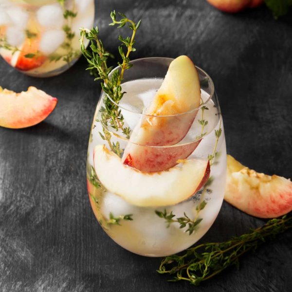 Fresh peach cocktail with ice, peach slices, and thyme on a dark background.