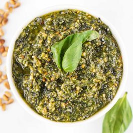 Square overhead photo of homemade basil pesto in a white bowl topped with fresh basil leaves and surrounded by toasted pine nuts.