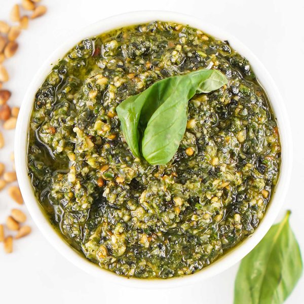 Square overhead photo of homemade basil pesto in a white bowl topped with fresh basil leaves and surrounded by toasted pine nuts.