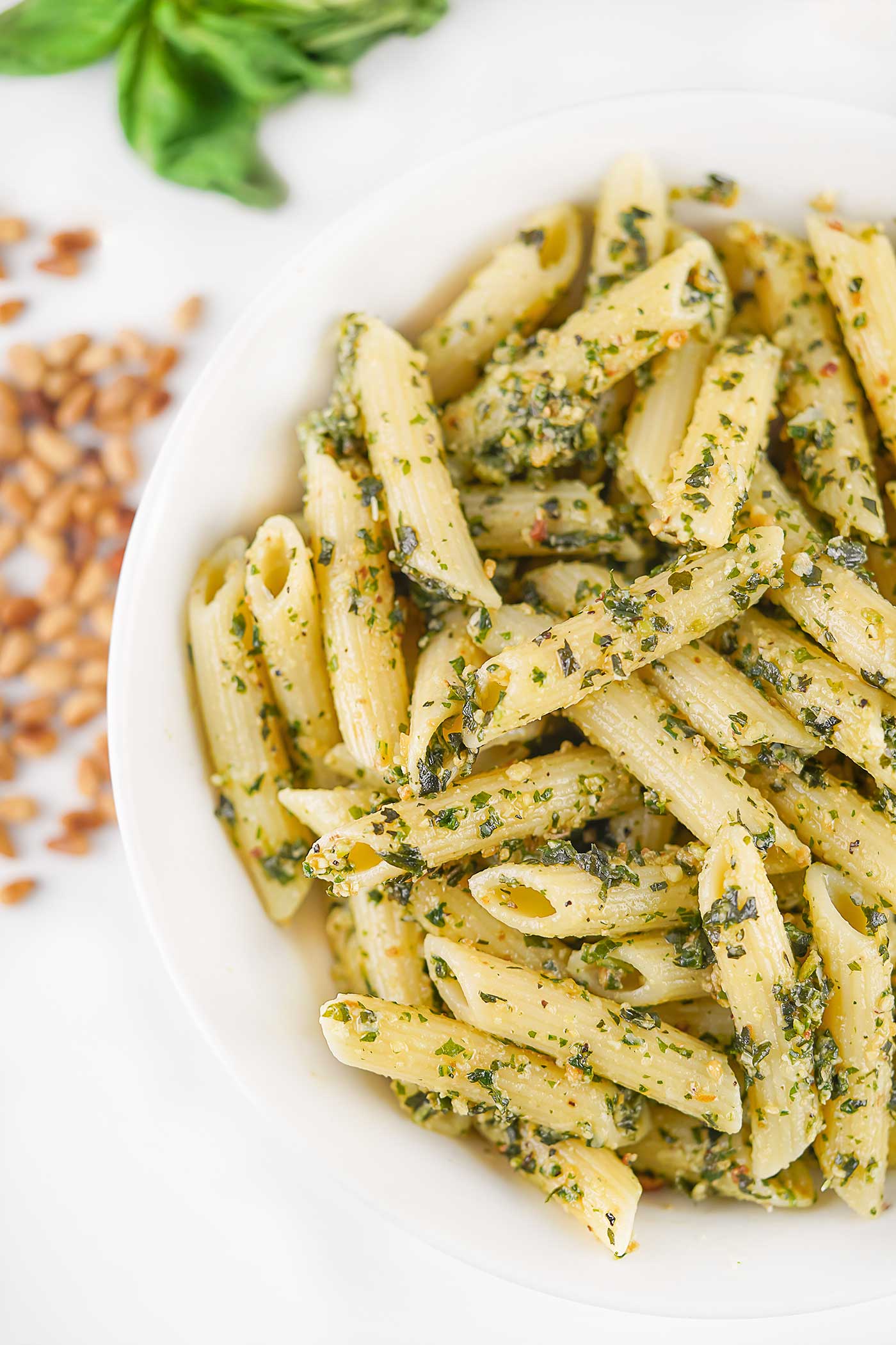 Penne pasta tossed with homemade basil pesto in a white bowl with fresh basil and pine nuts nearby.