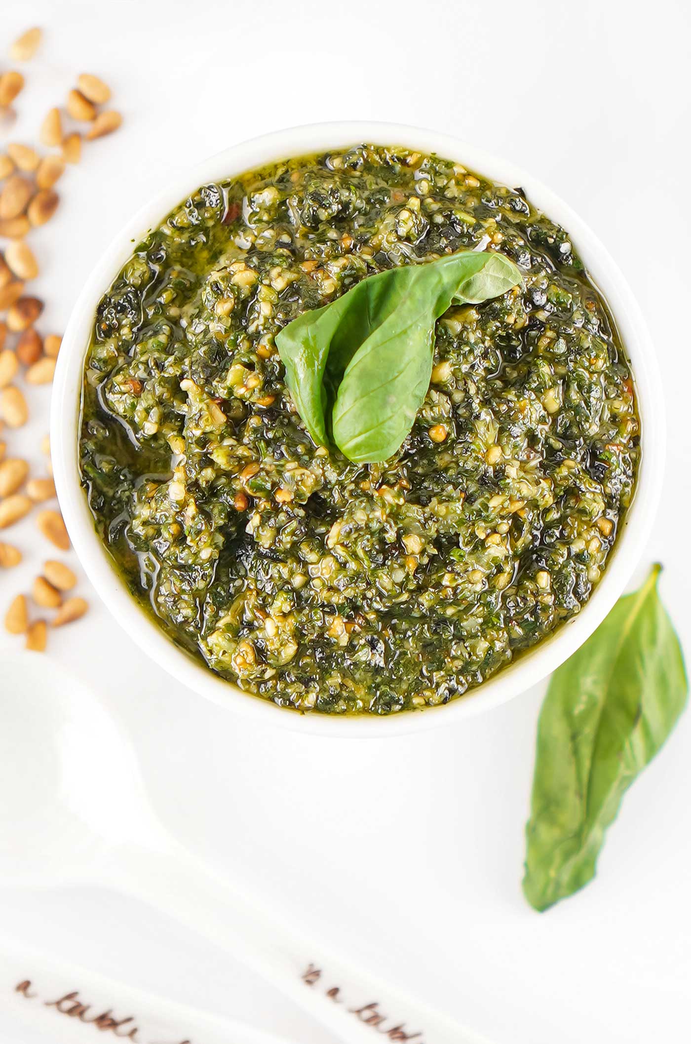 Overhead close-up of pesto in a white bowl topped with fresh basil leaves, with toasted pine nuts nearby.