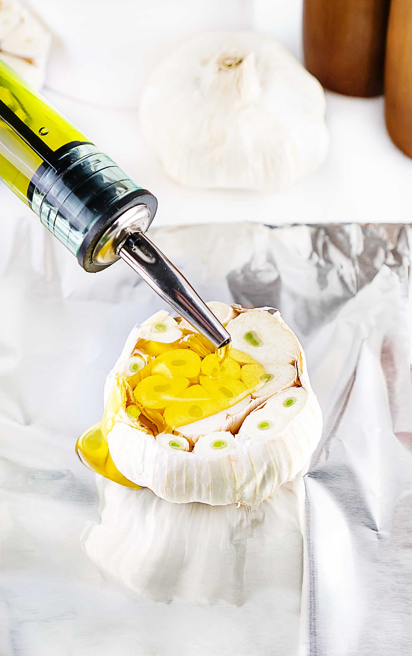 Olive oil being drizzled over the exposed cloves of a whole garlic bulb on aluminum foil.