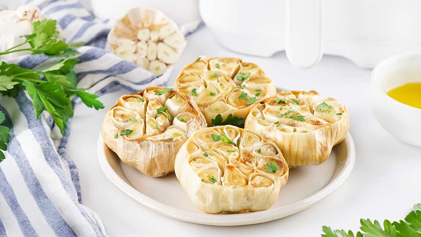 Roasted garlic bulbs on a plate with olive oil, parsley, and an air fryer in the background.