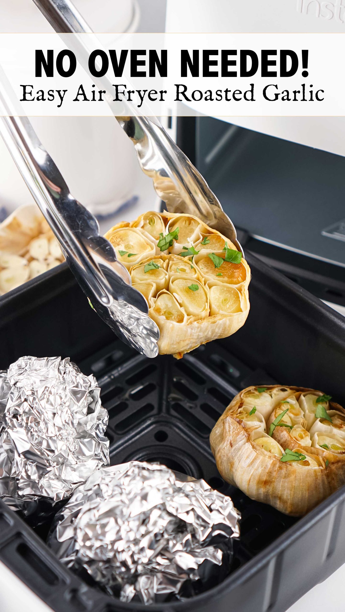 Kitchen tongs lifting a roasted garlic bulb from an air fryer basket with additional roasted and foil-wrapped bulbs inside.