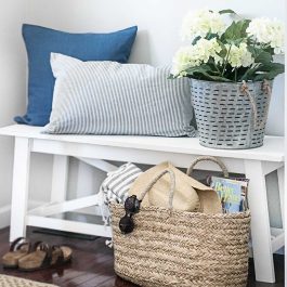 White entryway bench styled with blue and striped pillows, white hydrangeas, and a woven basket holding a sun hat, sunglasses, and magazines.