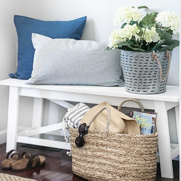 White entryway bench styled with blue and striped pillows, white hydrangeas, and a woven basket holding a sun hat, sunglasses, and magazines.