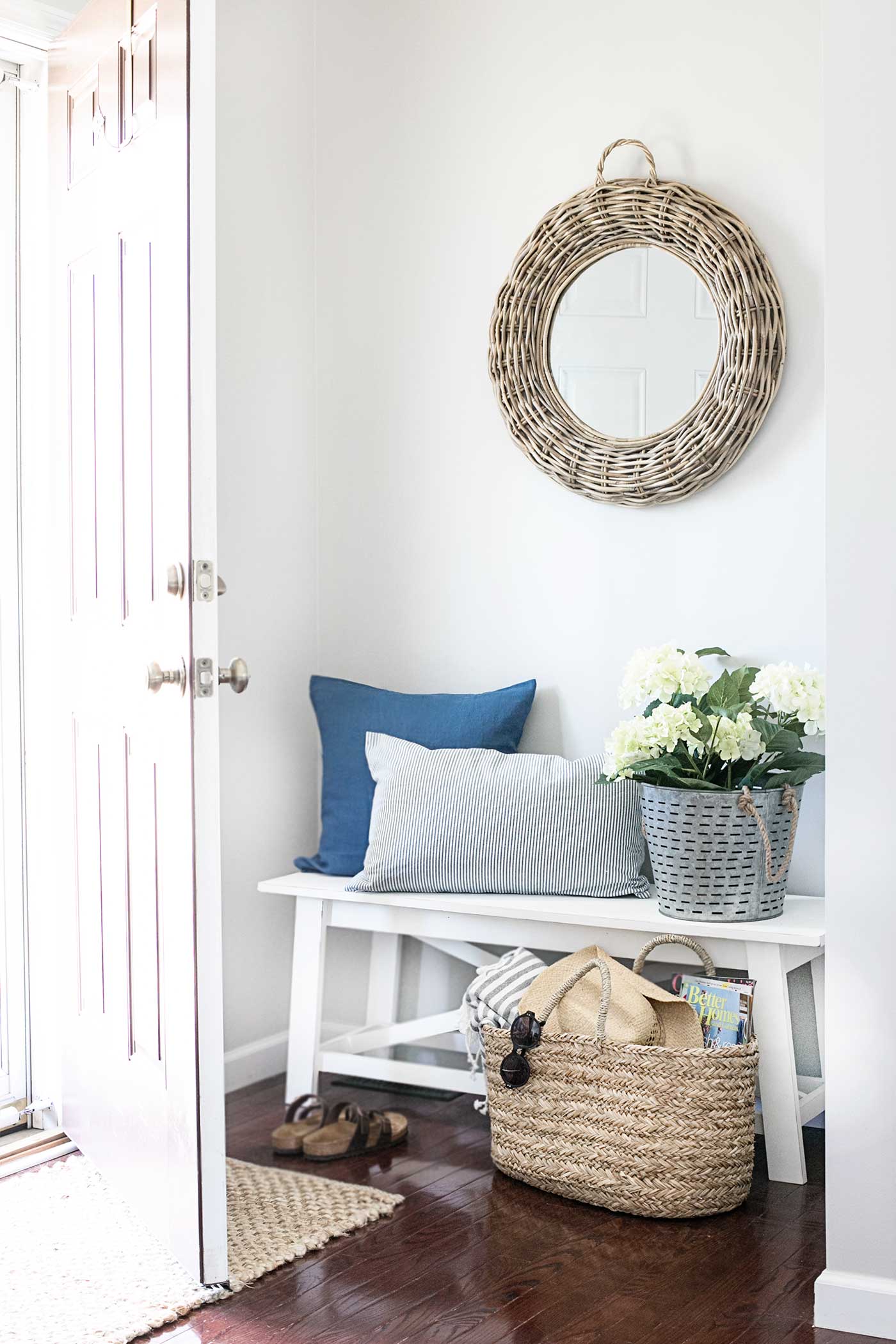Bright summer entryway with a white bench, blue and striped pillows, woven basket, white flowers, and round wicker mirror.