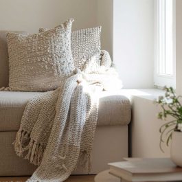 Sunlit neutral chair styled with textured cream pillows and a chunky throw beside a bright window and stacked books.