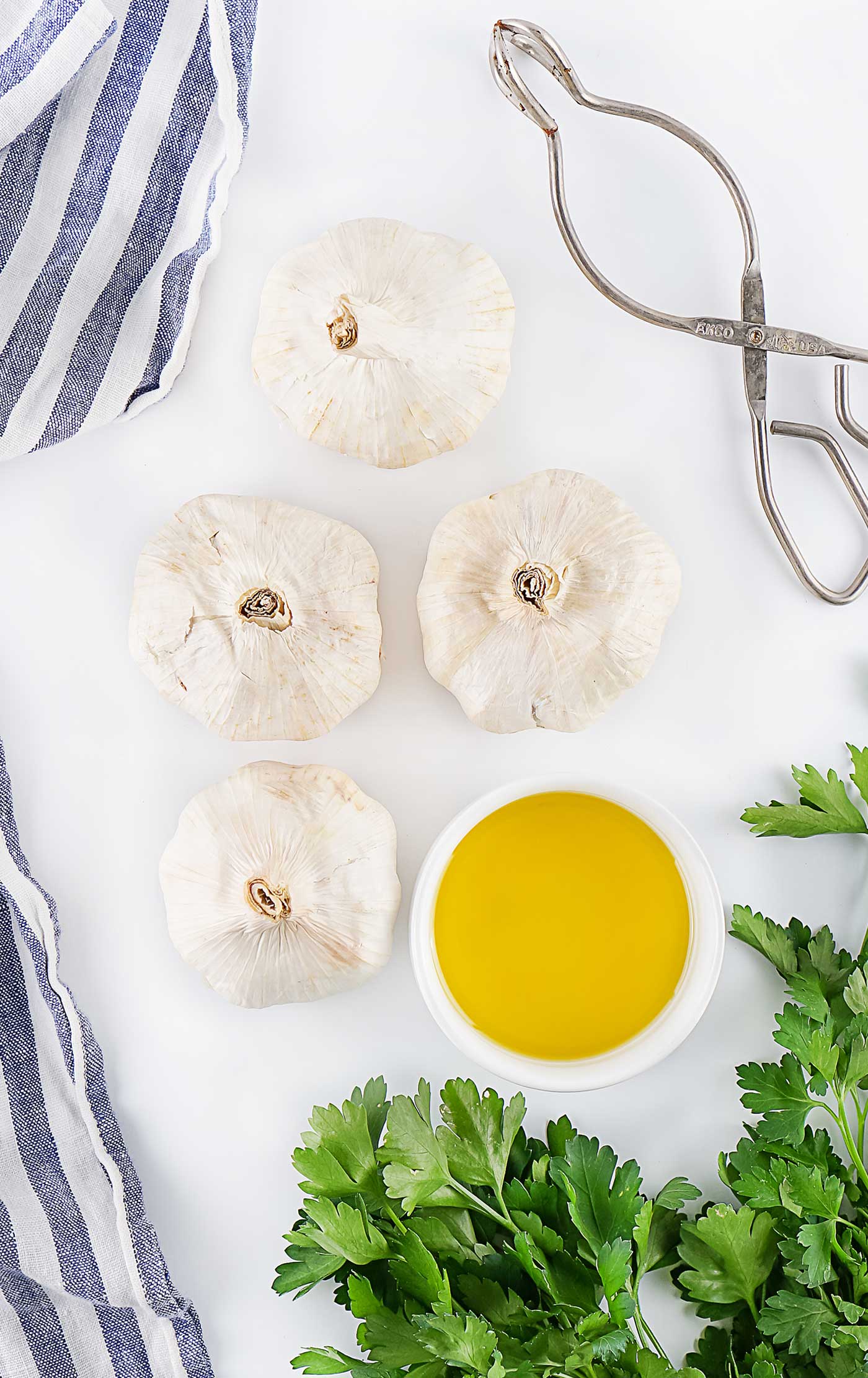 Whole garlic bulbs with a bowl of olive oil, fresh parsley, striped towel, and kitchen tongs.