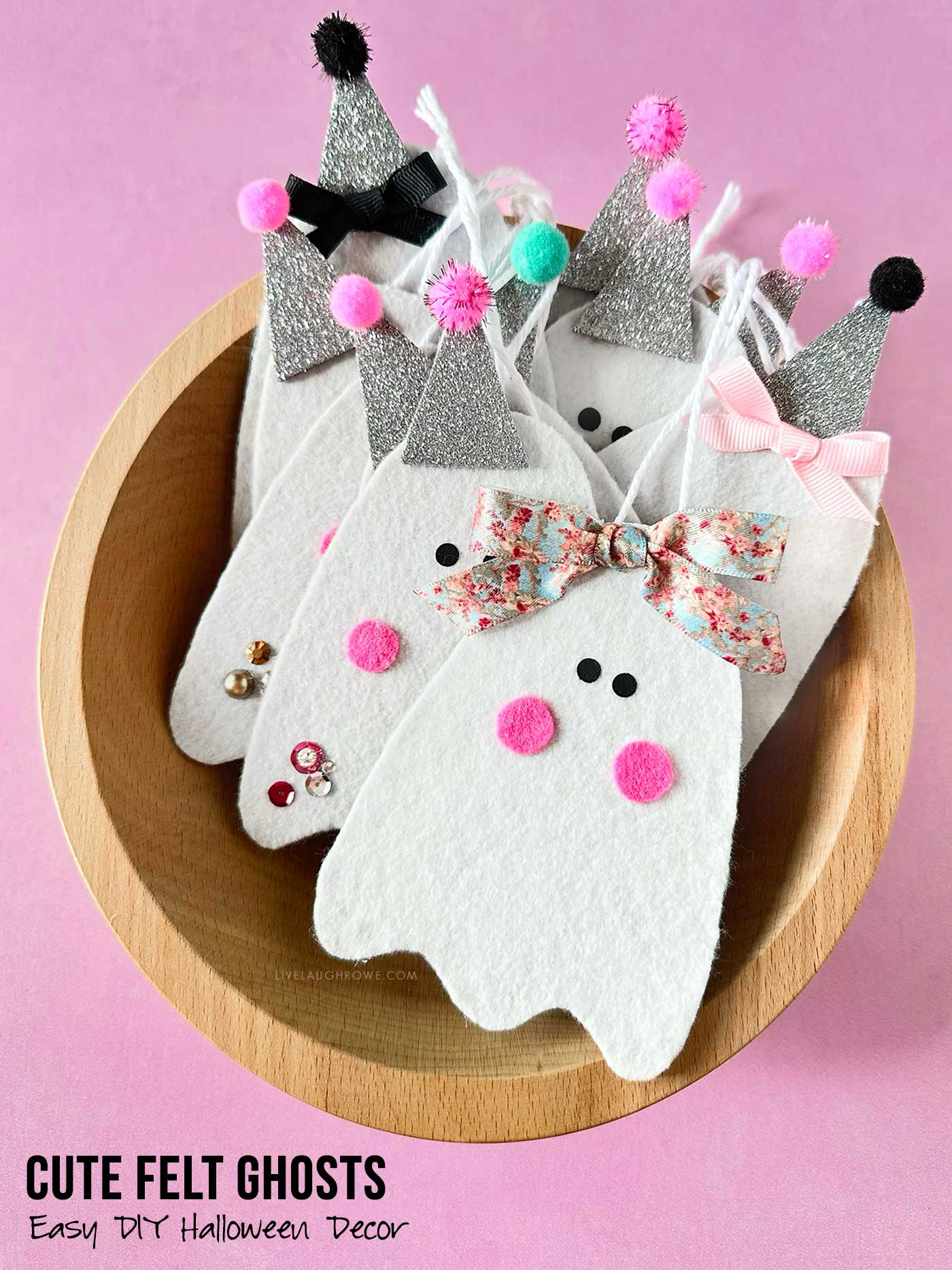 Cute handmade felt ghosts with glitter hats, bows, pom-poms, and embellishments displayed in a wooden bowl