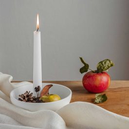 Red apple beside a lit white taper candle, pinecones, and neutral linen on a wooden table for a cozy early fall scene.