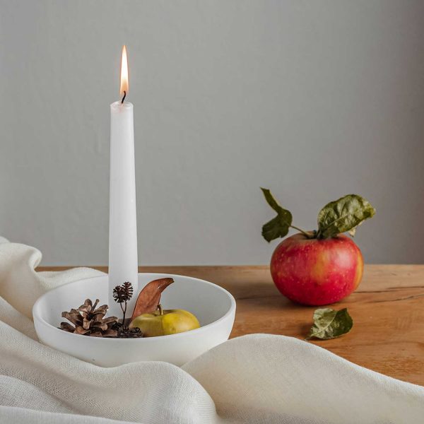 Red apple beside a lit white taper candle, pinecones, and neutral linen on a wooden table for a cozy early fall scene.
