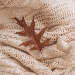 Brown autumn leaf resting on a cream chunky knit blanket.