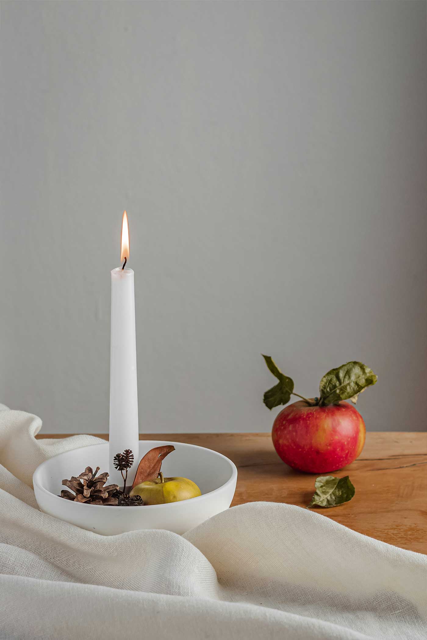 A cozy September still life with a red apple, white taper candle, pinecones, and soft neutral linen on a wooden table.