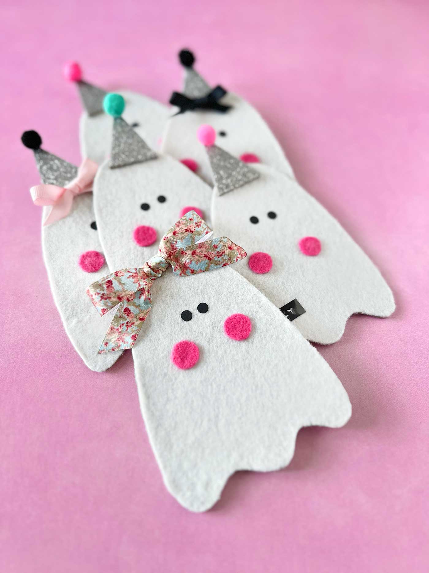 Group of handmade felt ghosts with glitter hats, pink cheeks, bows, and embellishments on a pink background