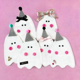 Group of handmade felt ghosts with glitter hats, bows, pink cheeks, and colorful embellishments on a pink background