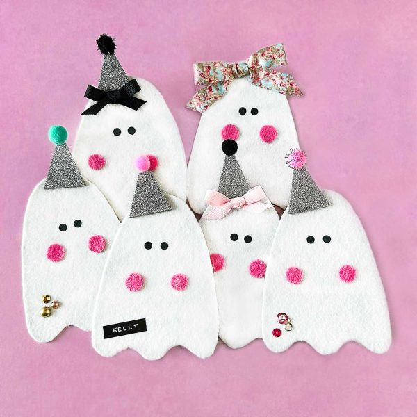 Group of handmade felt ghosts with glitter hats, bows, pink cheeks, and colorful embellishments on a pink background