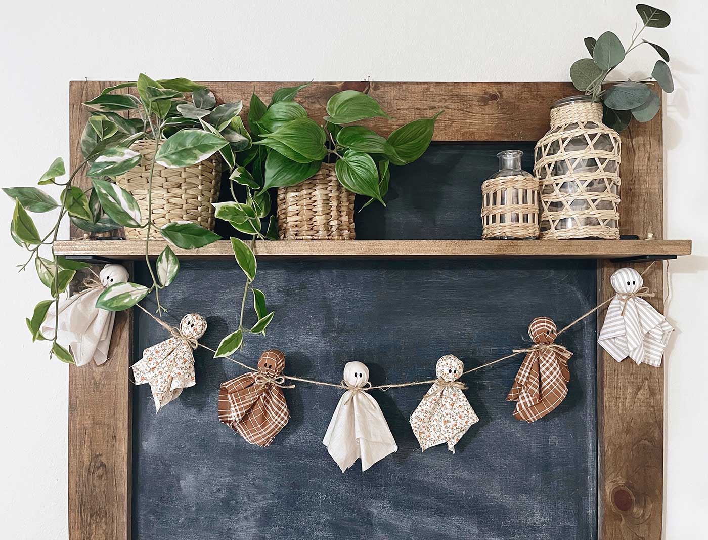 Handmade fabric ghost garland hanging across a large chalkboard with greenery and woven baskets