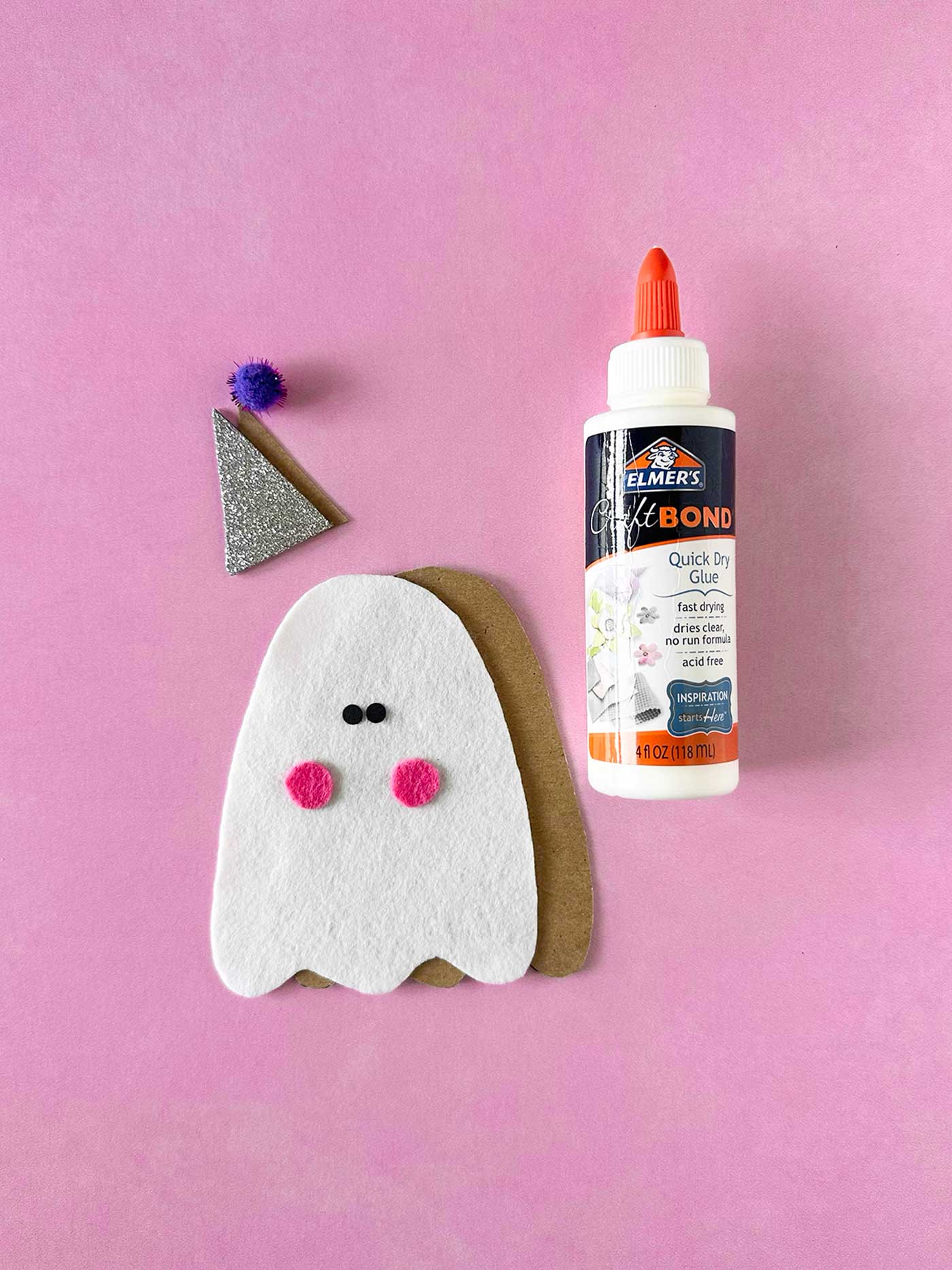 DIY felt ghost with pink cheeks beside glue, glitter hat, and pom-pom on a pink background