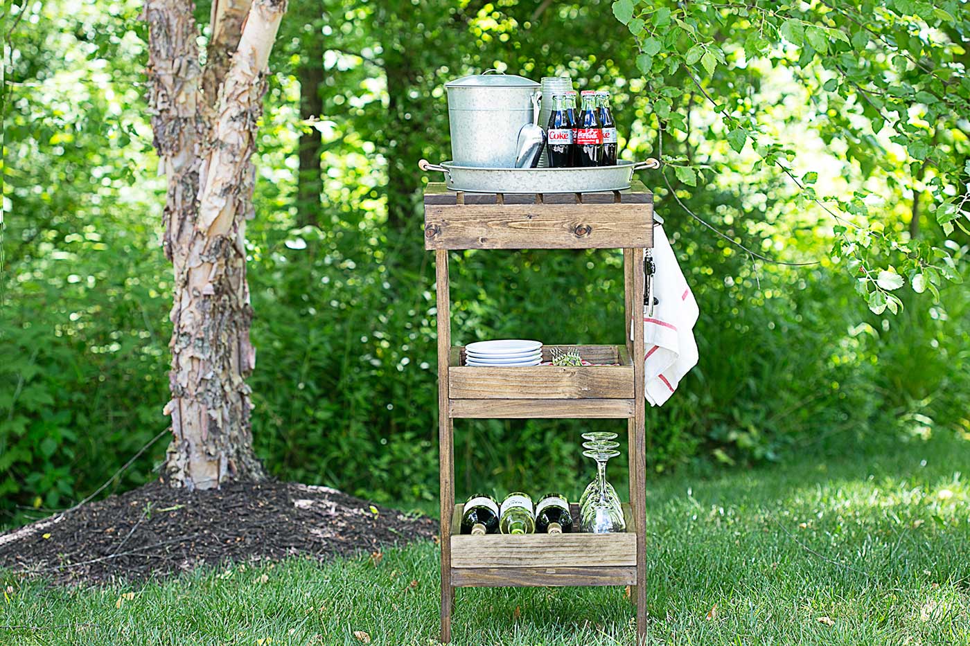 Wooden outdoor drink station styled with beverages and glasses beside a tree