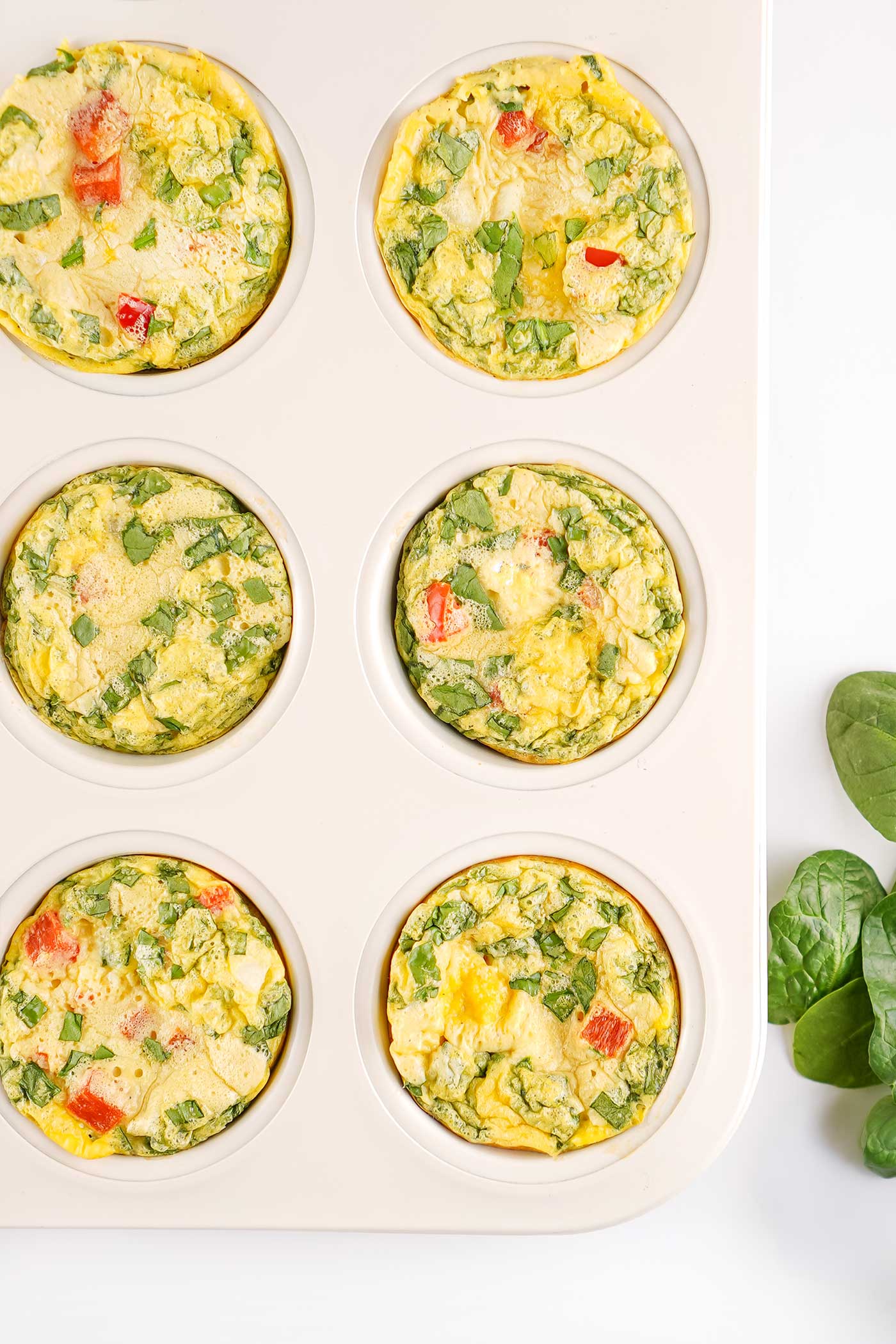 Six baked egg bites with spinach and red bell pepper in a white muffin pan
