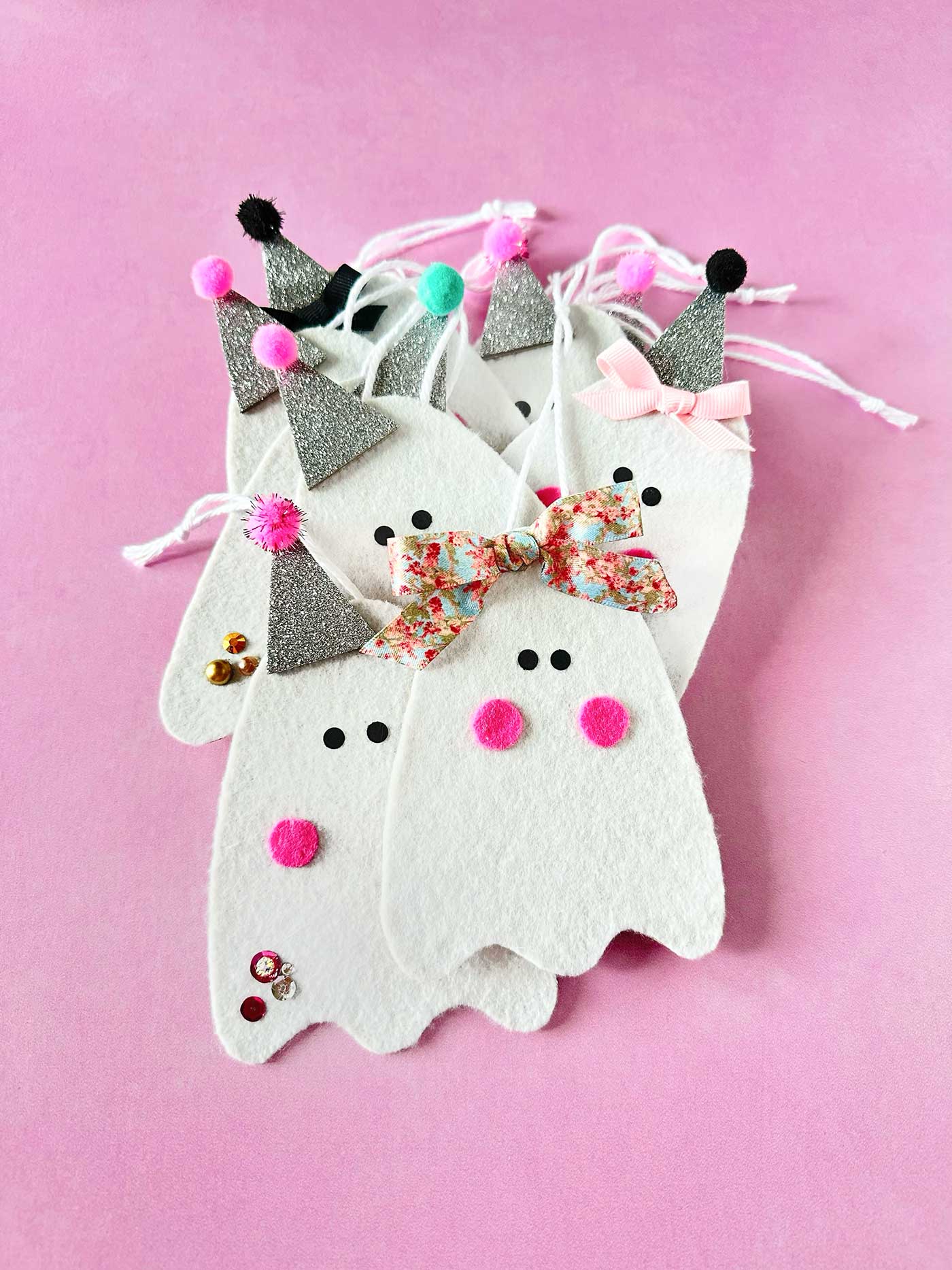 Handmade felt ghost ornaments with glitter hats, bows, pom-poms, and white hanging strings on a pink background
