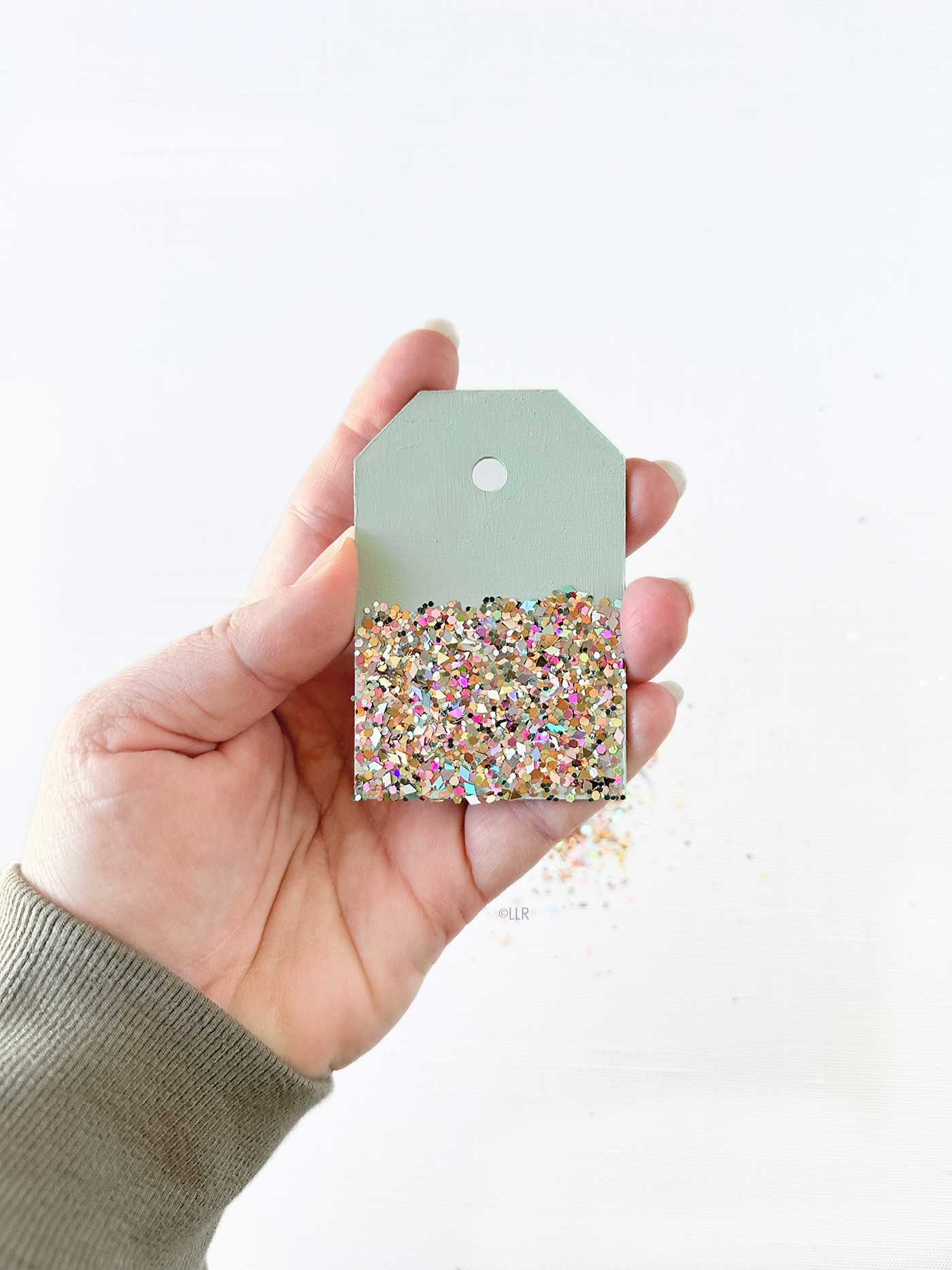 Hand holding a sage green wooden tag decorated with multicolored glitter