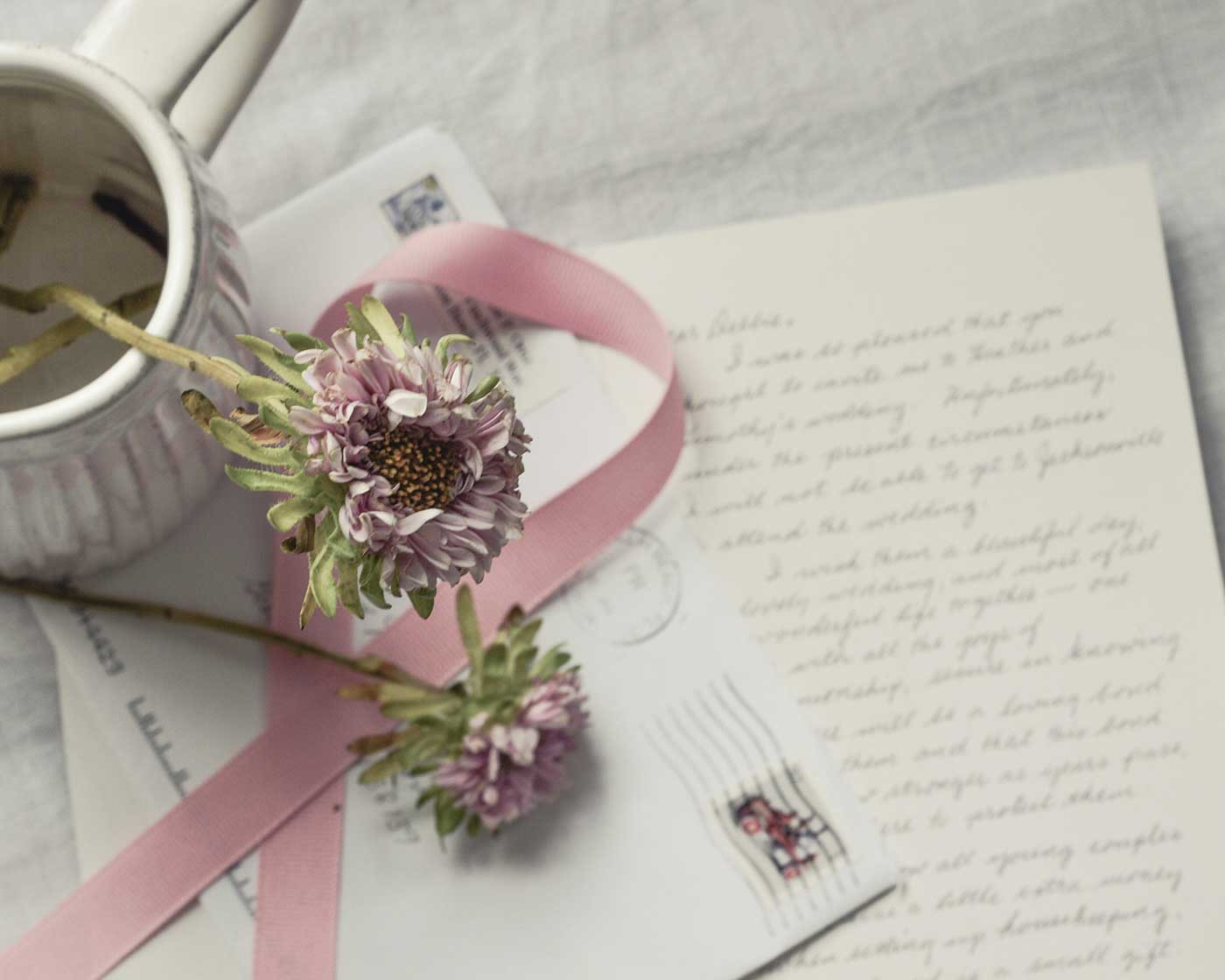 Handwritten letters and envelopes arranged with purple flowers, pink ribbon, and a ceramic pitcher.